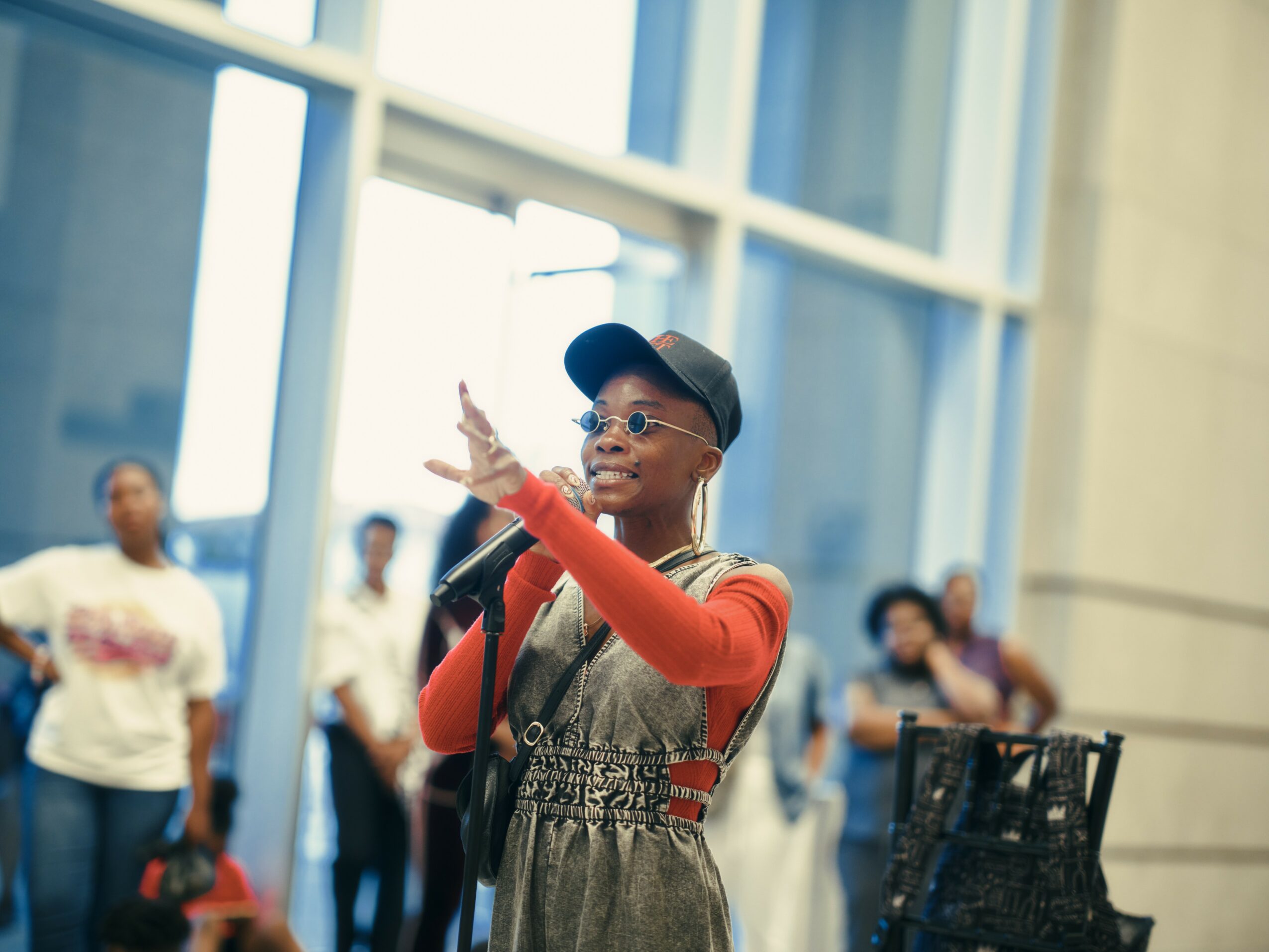 Young person speaking into a microphone, gesturing with hand, in a modern indoor space with large windows, group of people in background.