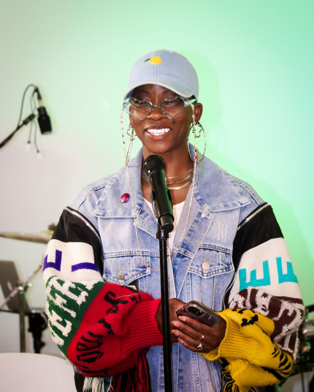 Person smiling, wearing a white cap, glasses, denim jacket, colorful sleeves, and holding a phone, standing in front of a microphone.