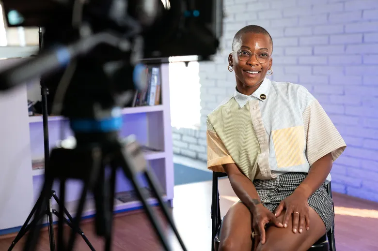 Woman smiling and sitting in front of camera during a video shoot, with a white brick wall background.