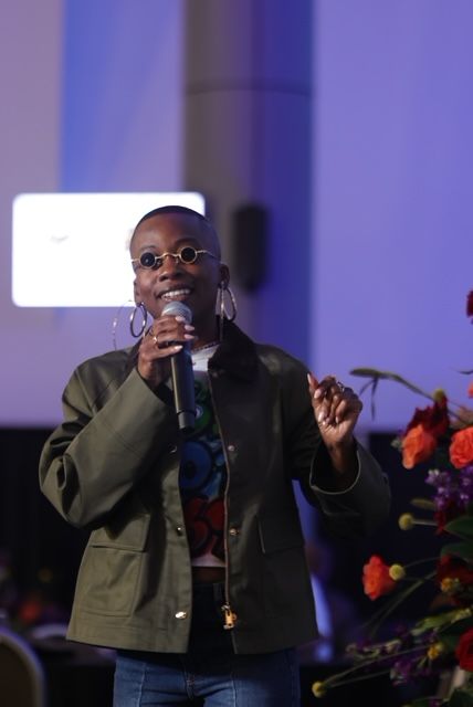 Person speaking into a microphone at an event, with a purple background and flowers in the foreground.