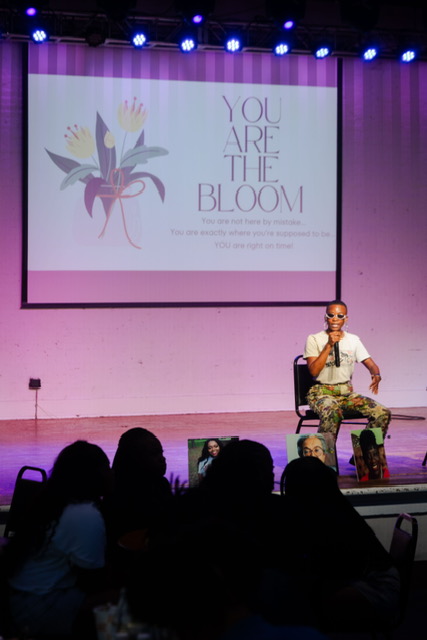 Person sitting on stage with a microphone, audience in foreground, large screen behind displaying a flower and text.