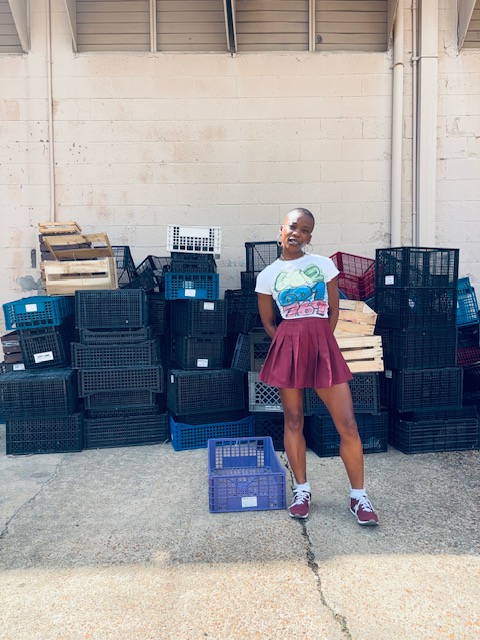 Person standing outdoors in front of stacked black and blue crates, wearing a white t-shirt and maroon skirt.
