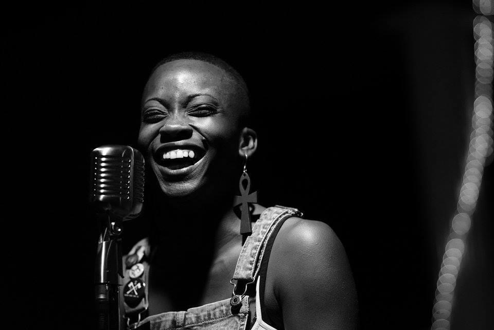 A woman laughing with a microphone, wearing earrings and a sleeveless top, in black and white.