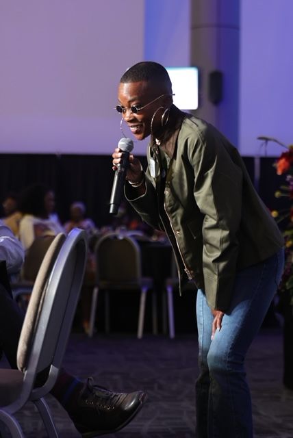 Person speaking into a microphone at an indoor event, with audience and chairs visible in the background.