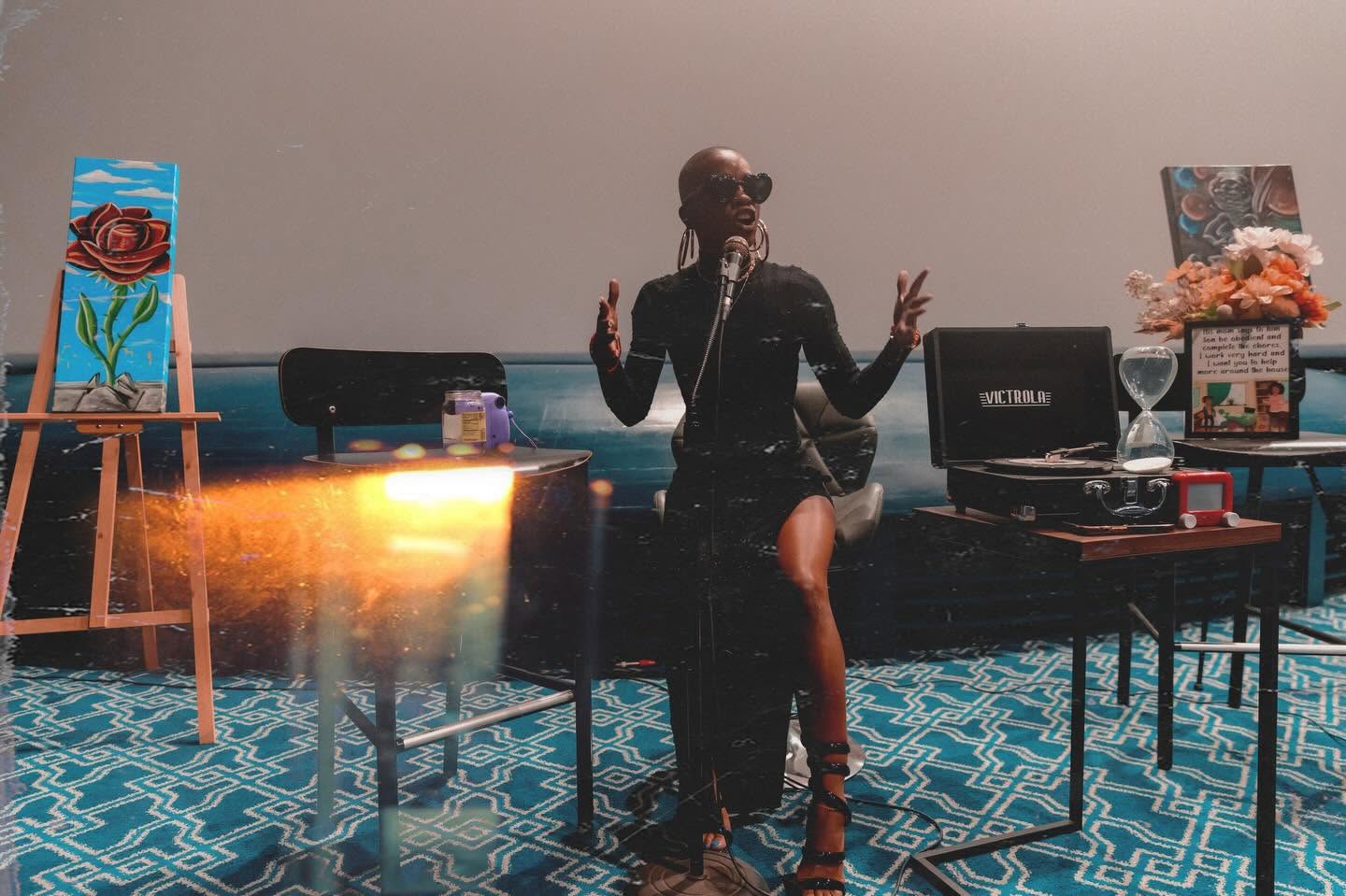 Person sitting on a bench with a microphone, surrounded by artwork and equipment, in a room with patterned blue carpet.