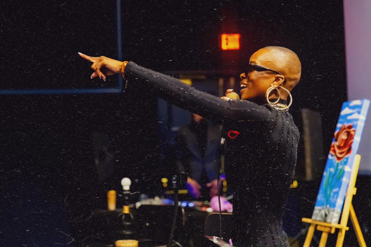 Bald woman with glasses and large earrings points to the left in a dark room with colorful artwork and equipment.