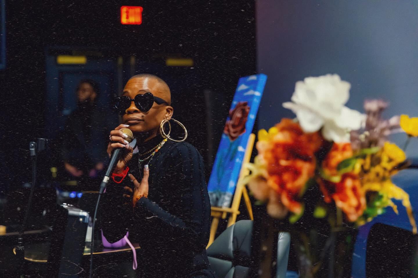 Person wearing sunglasses and earrings speaking into a microphone at an indoor event, with a floral arrangement nearby.