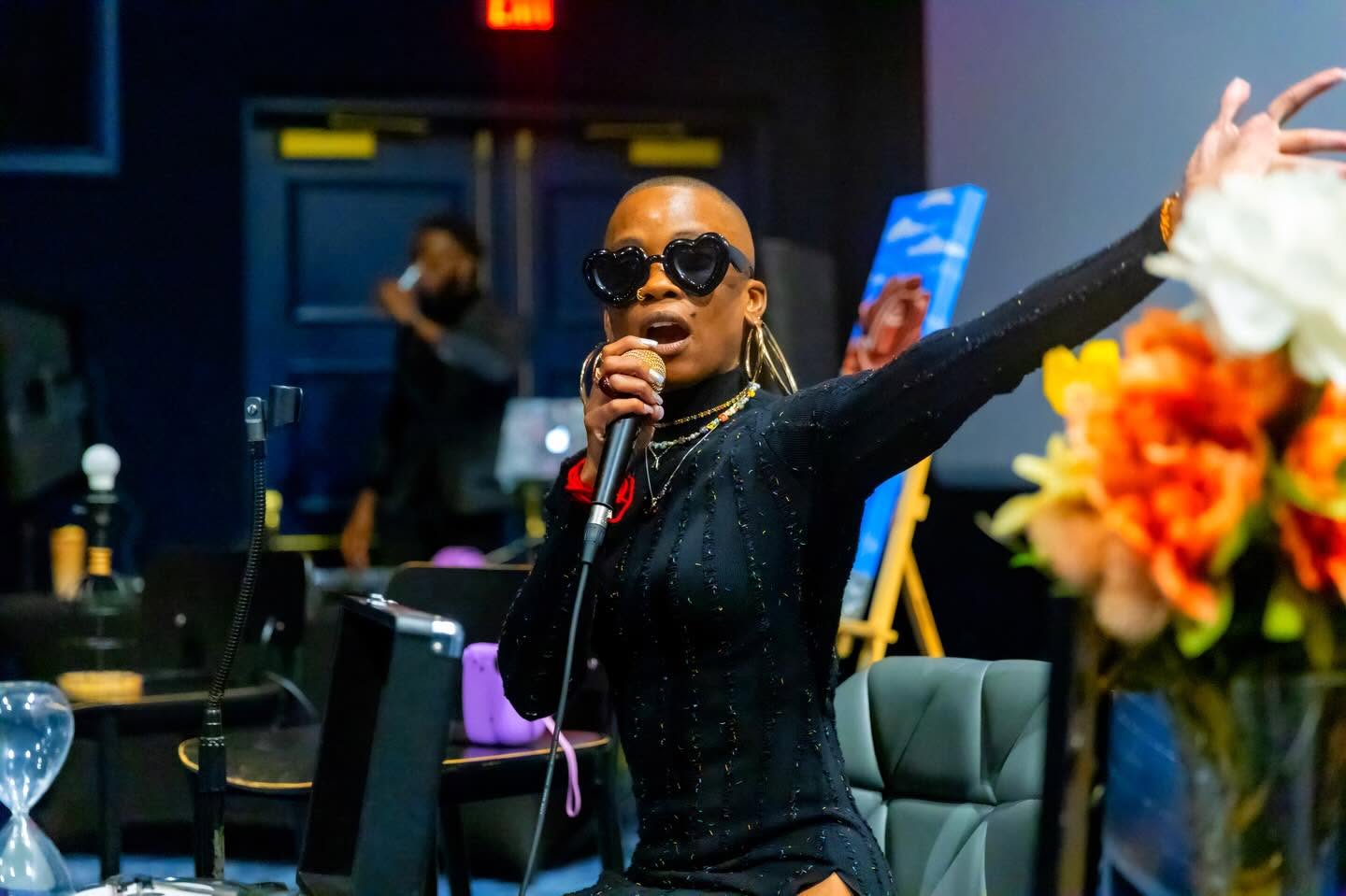 Woman singing into microphone with sunglasses, in a room with tables and chairs, and a flower arrangement in foreground.