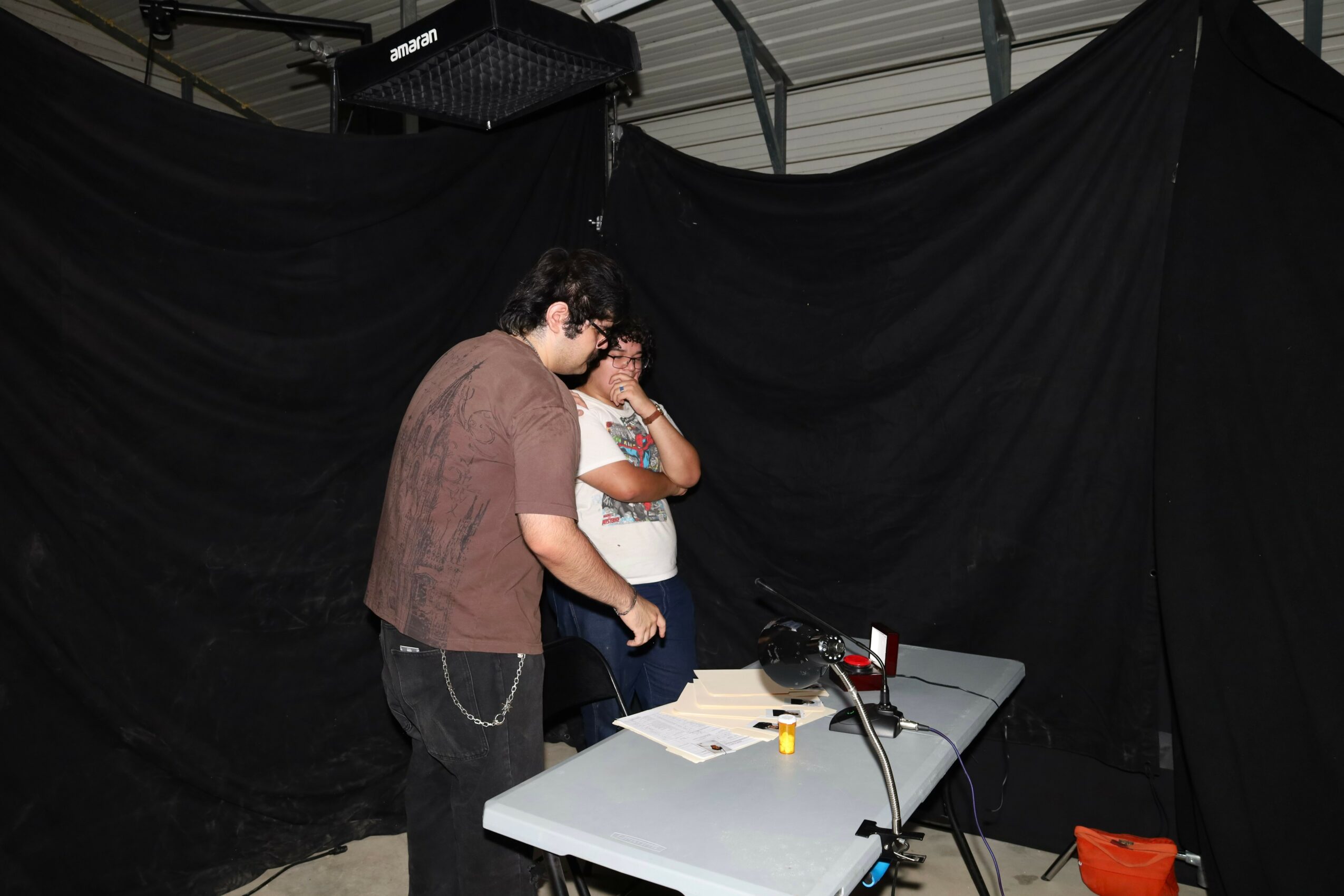 Two people standing at a table with papers and equipment, in front of a black backdrop, indoors.