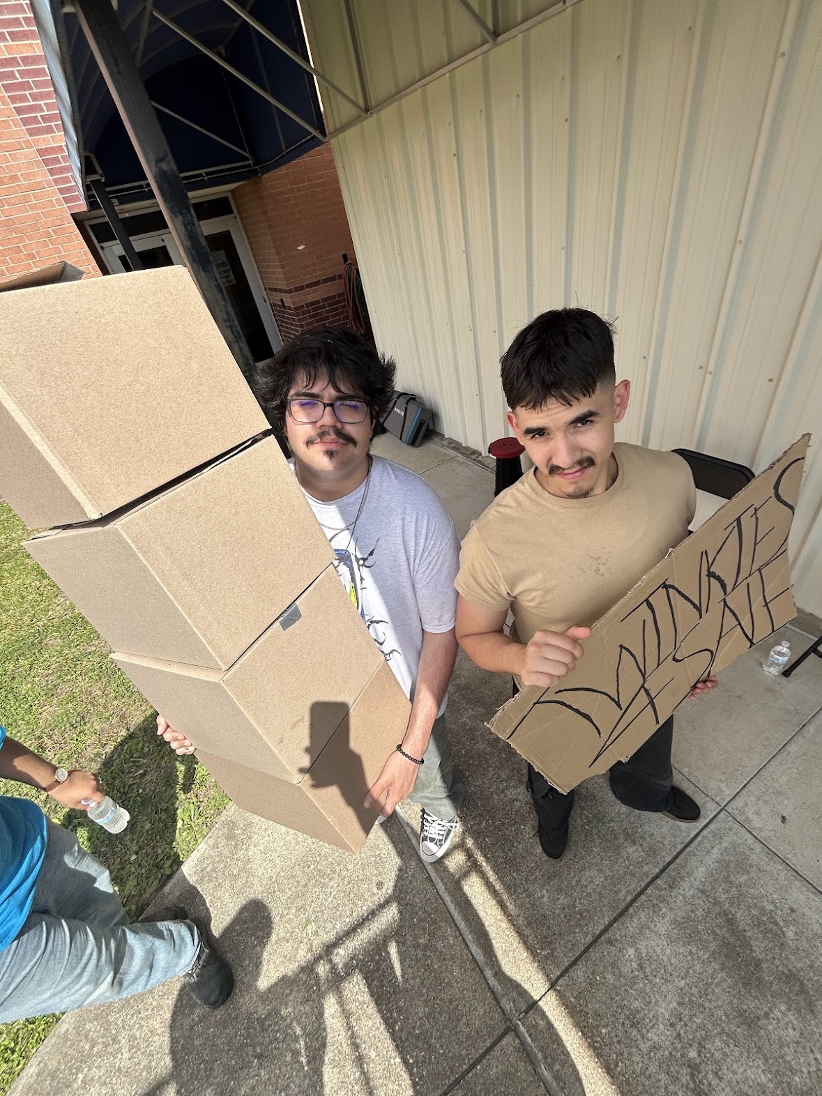 Two young men holding cardboard signs, standing outside near a building with a metal wall, smiling at the camera.