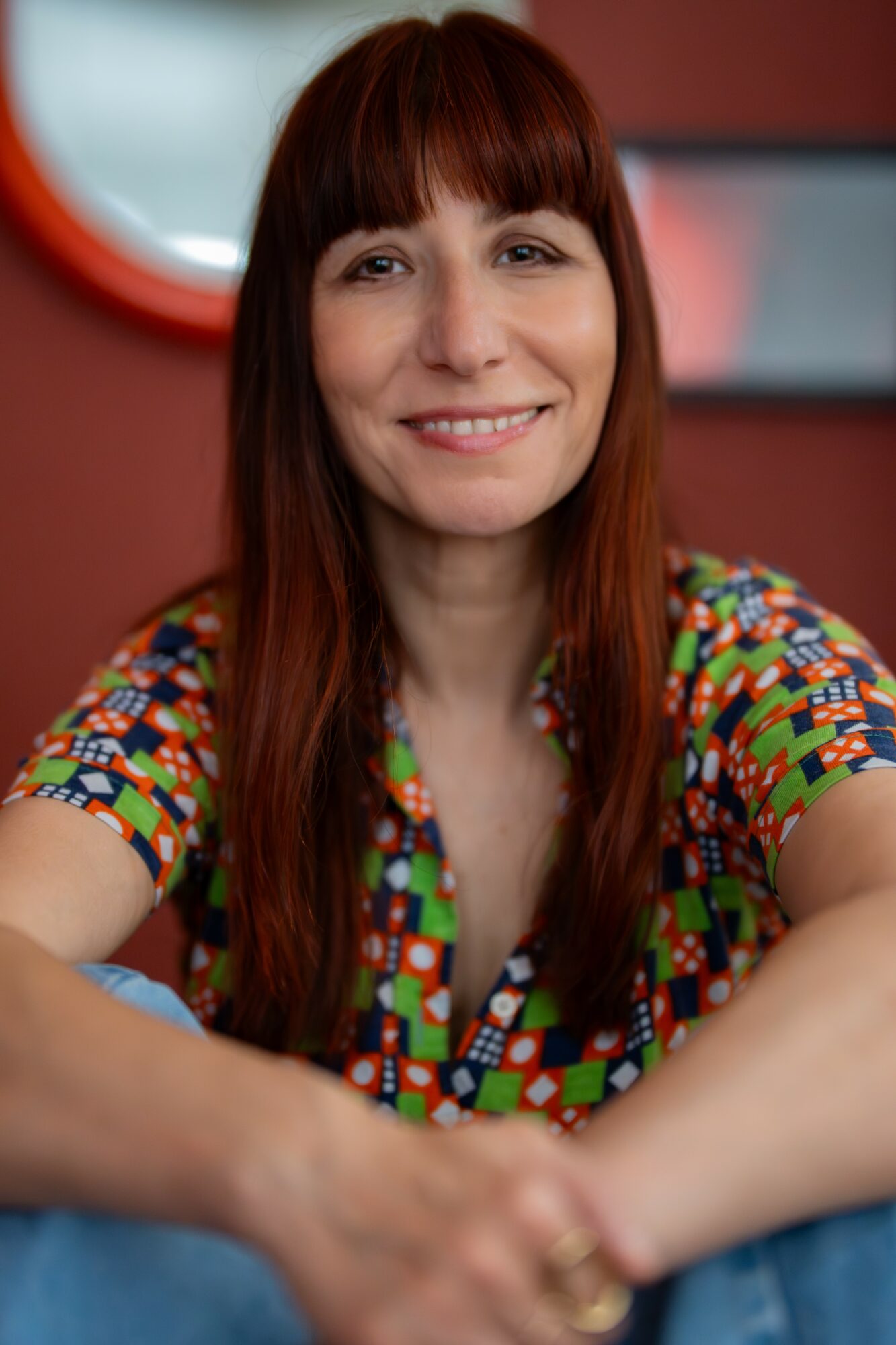 Woman with long red hair smiling, wearing a colorful patterned shirt, sitting indoors with a red wall and mirror behind her.
