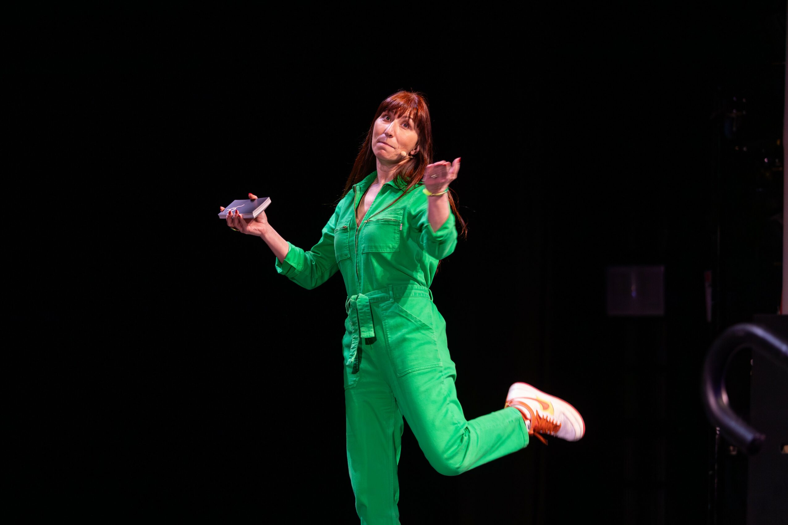 Woman in green jumpsuit on stage, holding a microphone, with one leg raised and hand extended, against a black background.