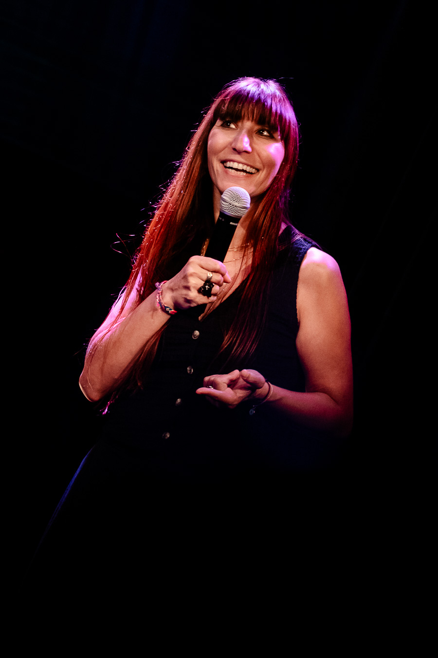 Woman with long hair holding a microphone, smiling on stage against a dark background.