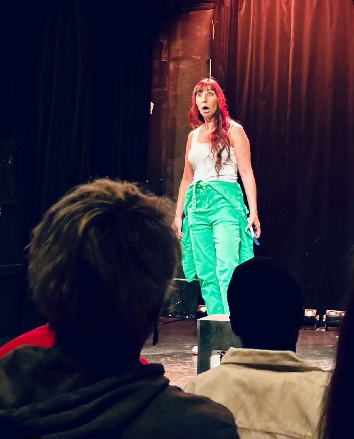 A woman with long hair and a white top stands on stage with a black curtain background, performing or speaking.