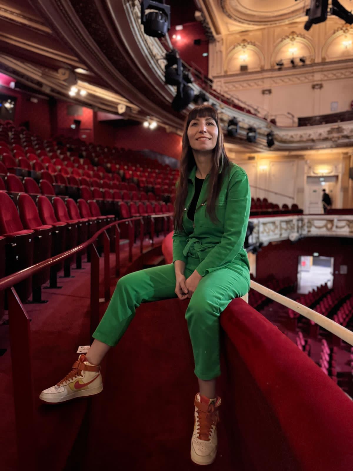 Woman in green outfit sitting on a red theater balcony, with ornate interior and empty red seats in background.