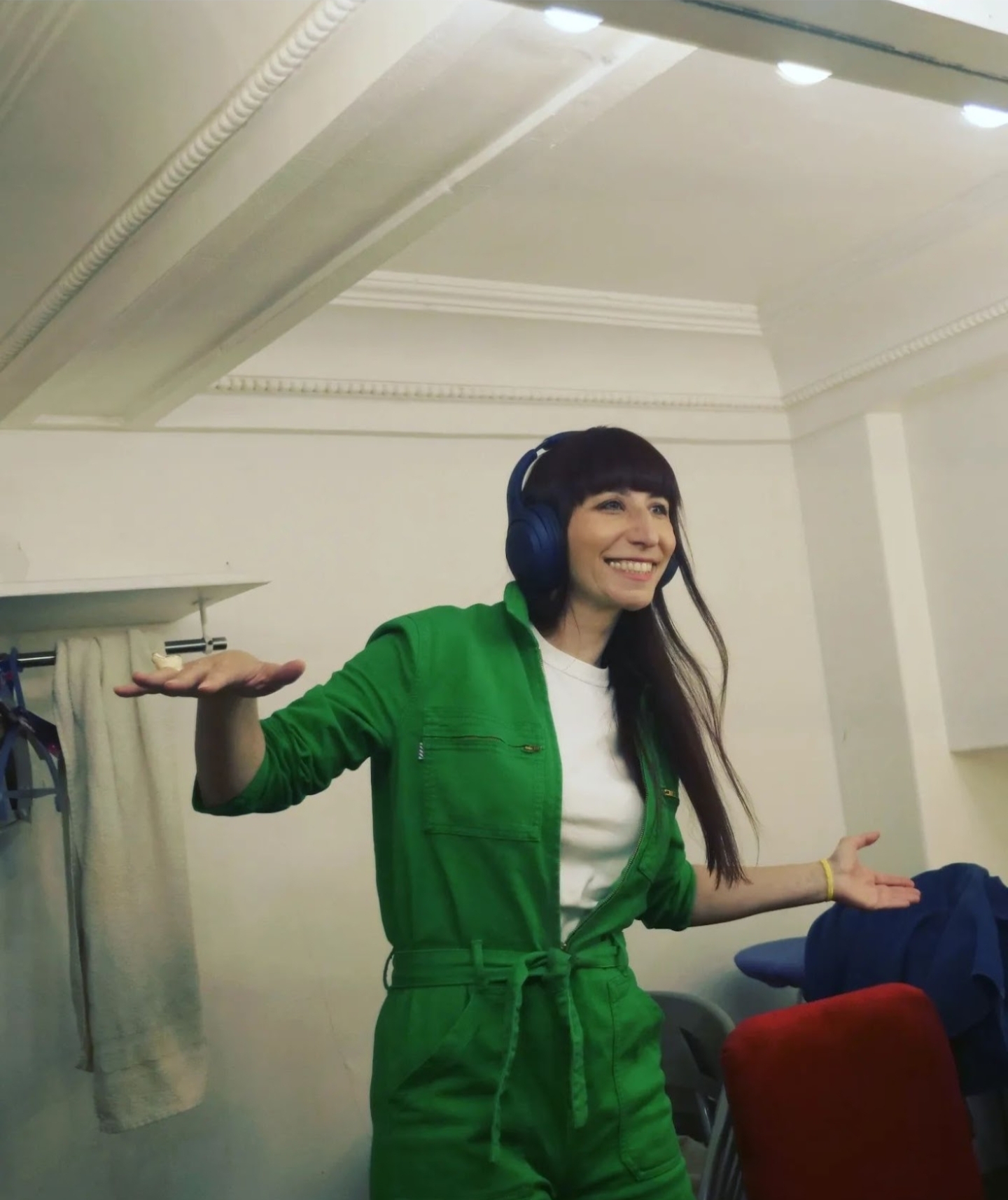 Woman wearing green jumpsuit and headphones, smiling, standing indoors with her arms outstretched.