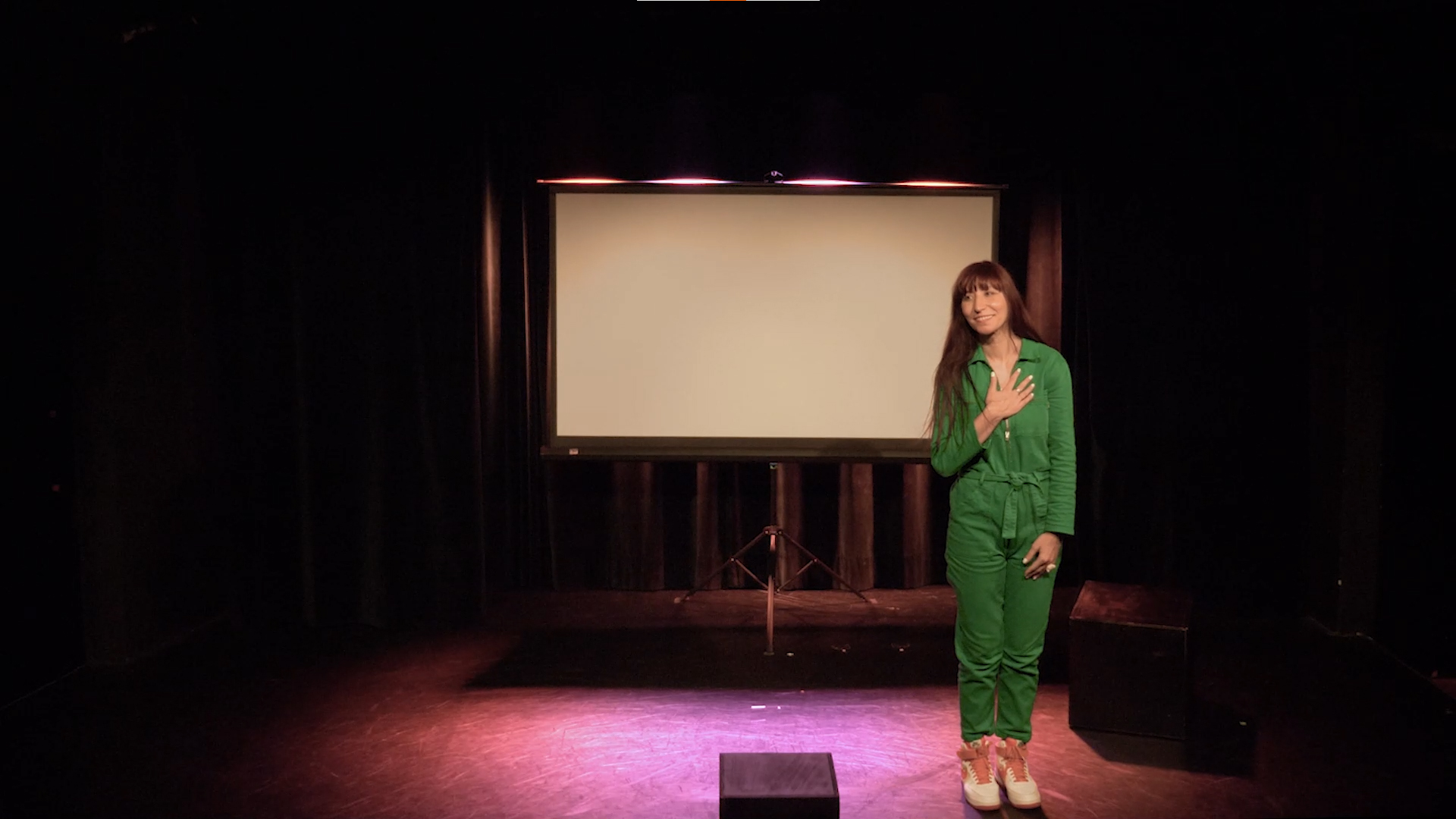 A woman in green outfit stands on stage with a large blank screen behind her, in a dark theater setting.