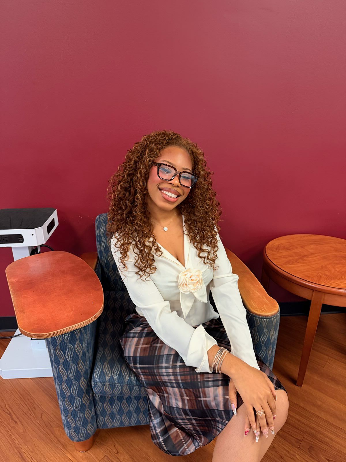 Woman with curly hair and glasses sitting on a chair, smiling, in a room with pink wall and wooden furniture.