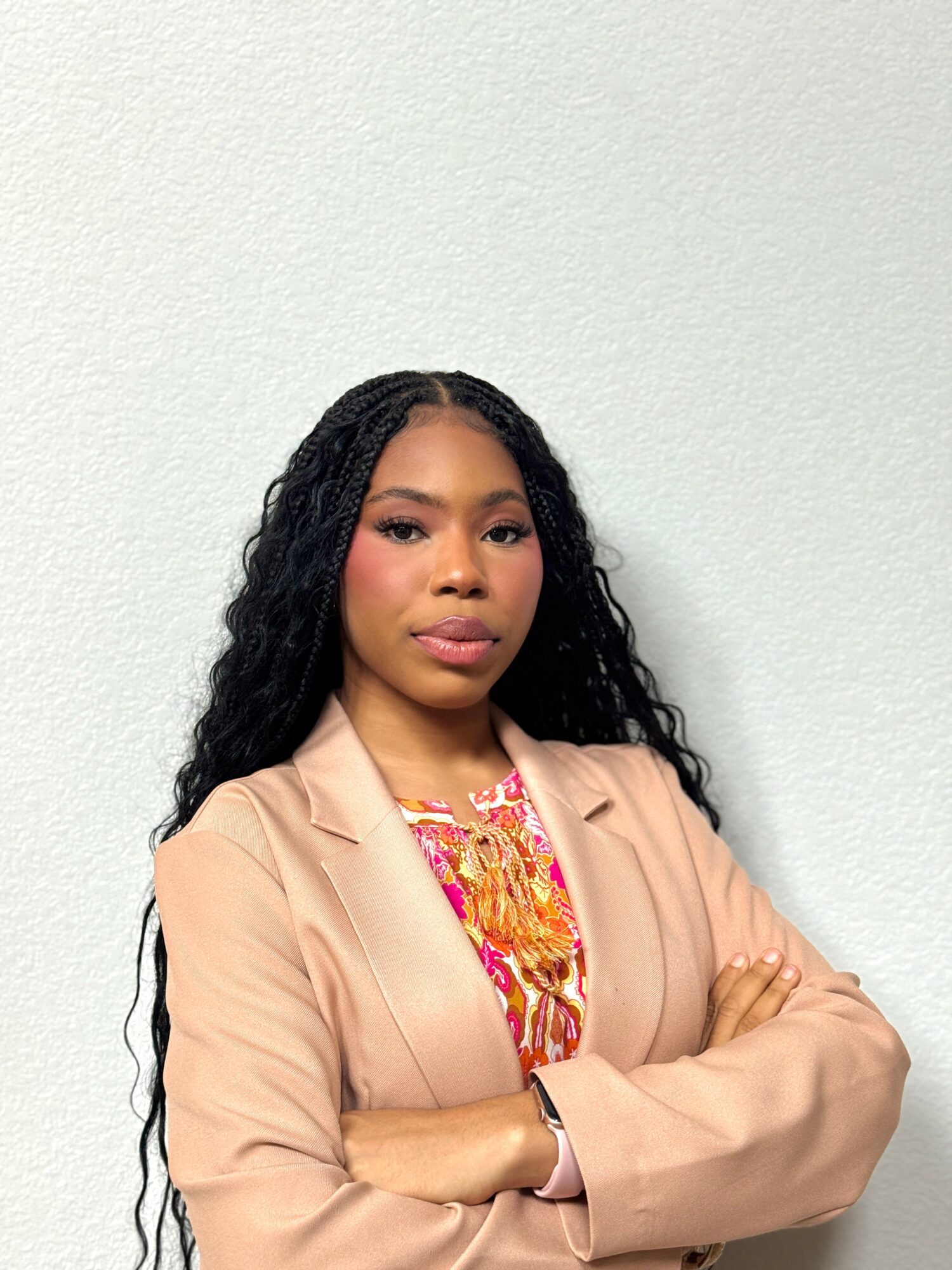 Woman with long curly hair wearing a beige blazer and colorful top, standing with arms crossed against a plain light background.