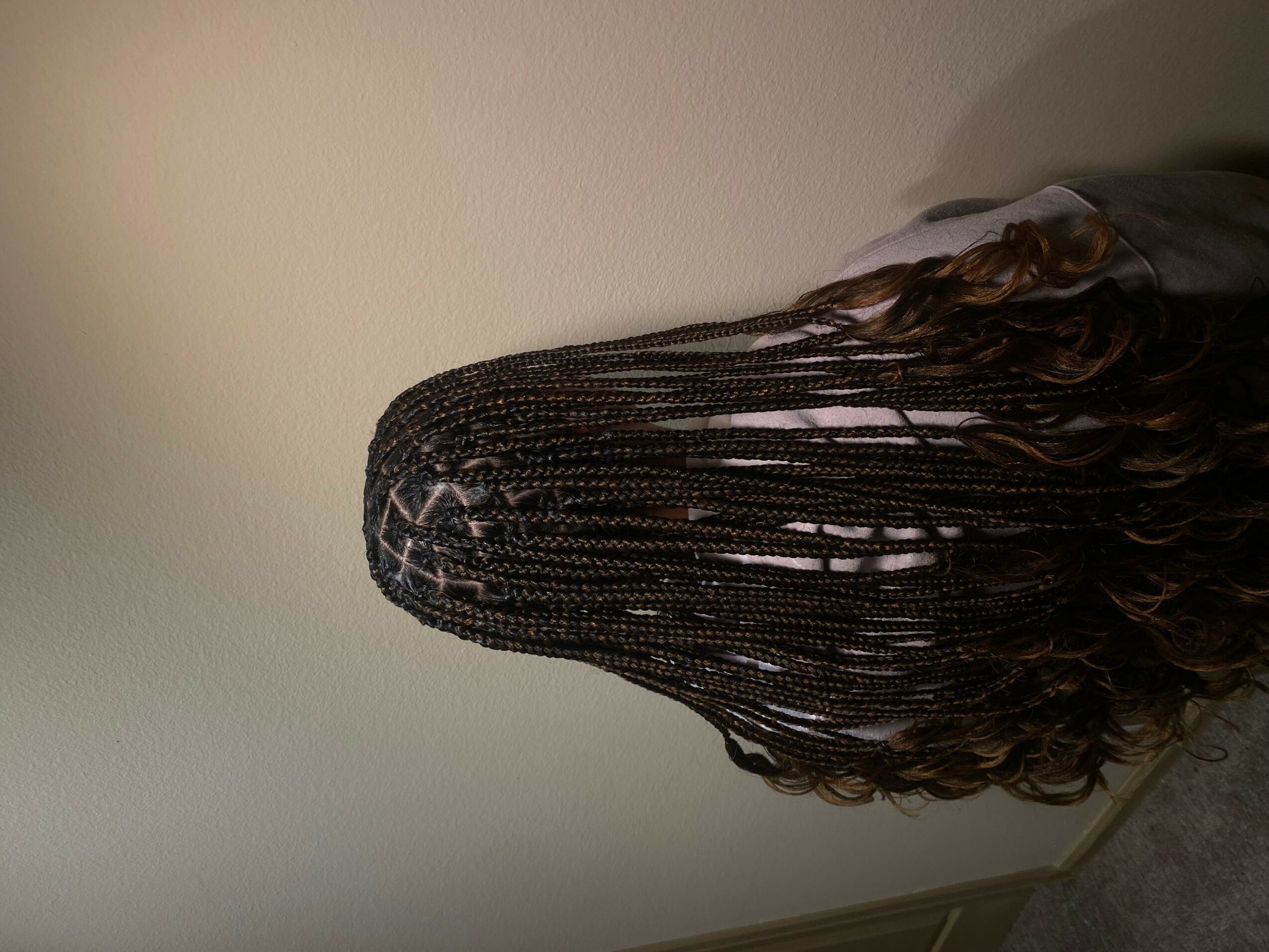 Person with long braided hair facing a plain wall, head turned away from the camera.