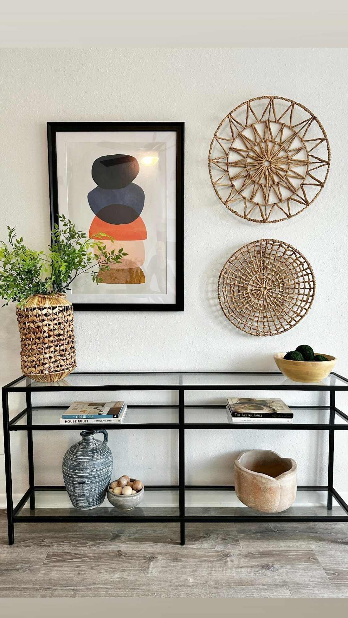Decorative wall with framed artwork, woven wall hangings, and a black metal console table with plants, vases, and bowls.