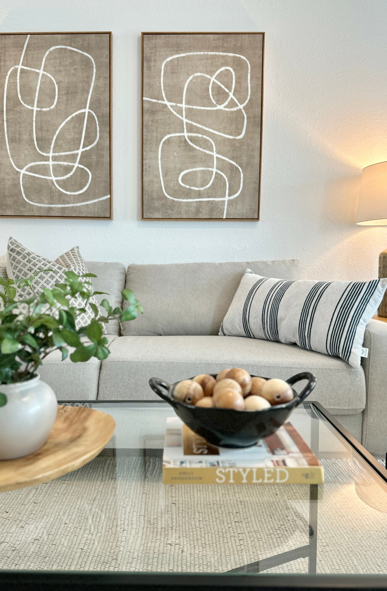 Living room with abstract line art paintings, a light-colored sofa with striped pillows, a glass coffee table, and a lamp.