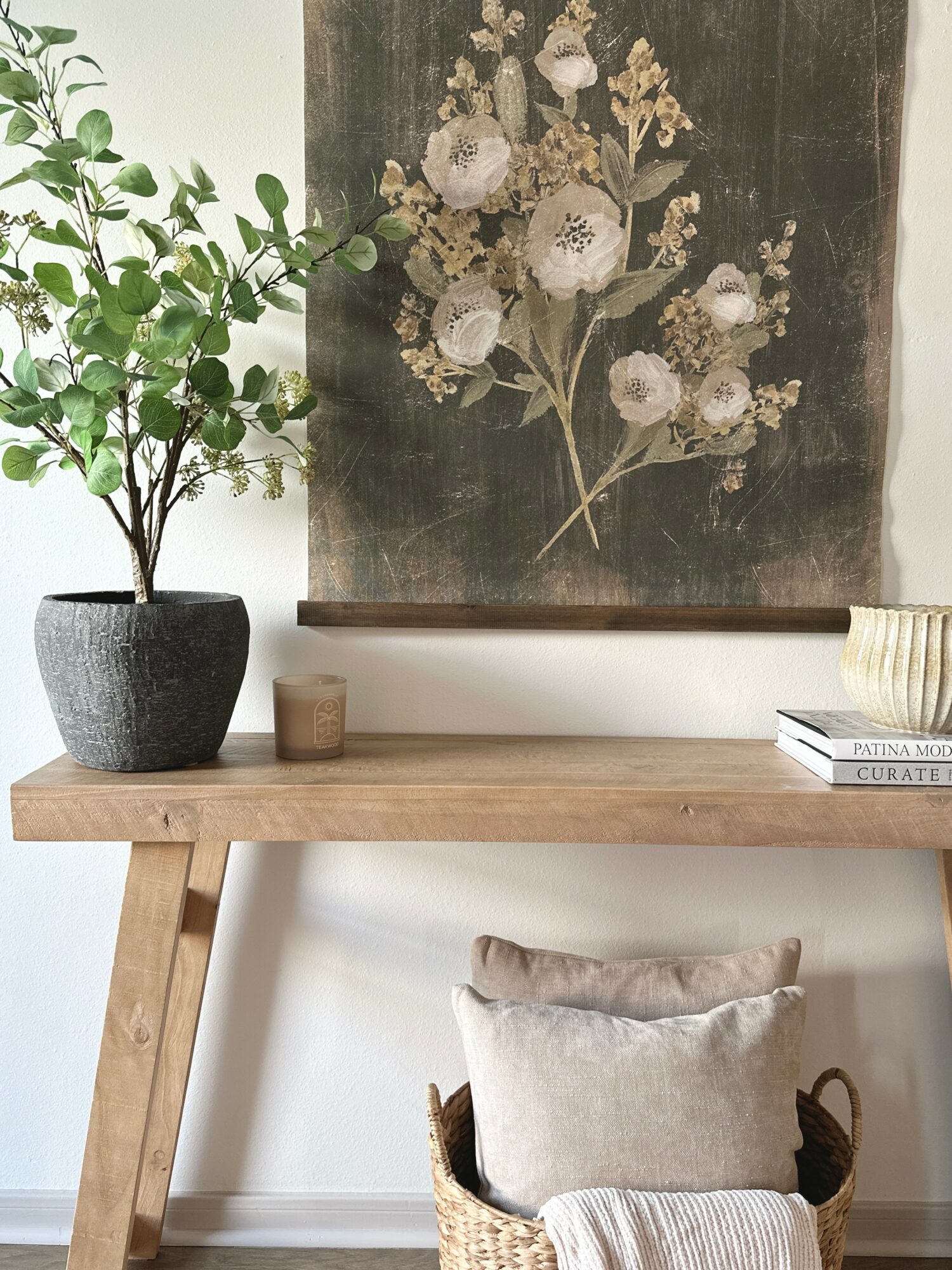Interior scene with a wooden table, potted plant, framed artwork, and a cushion on a woven basket, against a white wall.