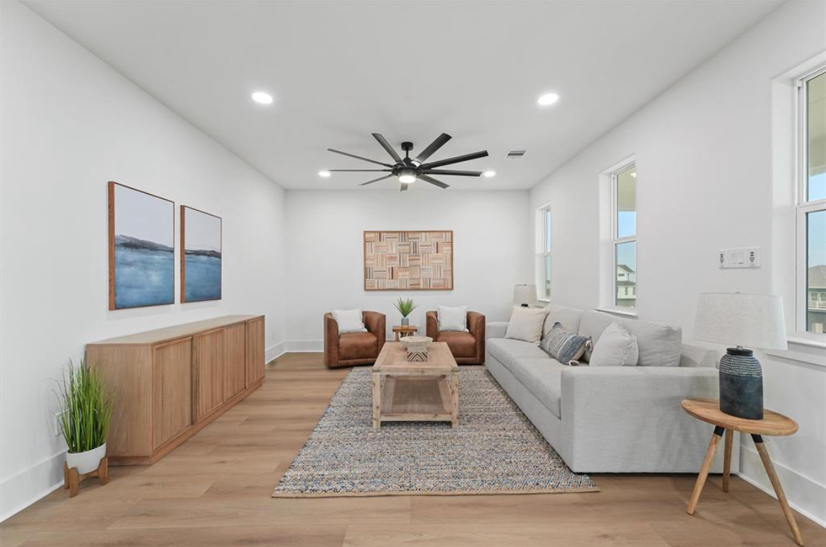 Living room with white walls, wooden floor, sofa, armchairs, coffee table, and large windows, decorated with artwork and plants.