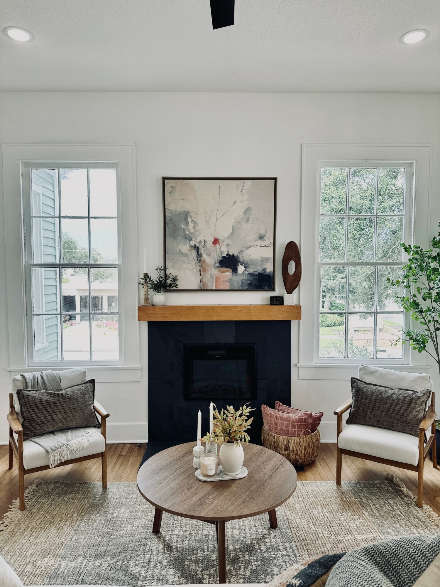 Living room with two windows, a fireplace, two chairs, a round table, and a painting above the fireplace.