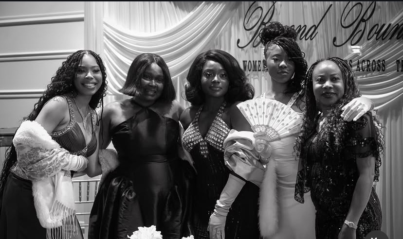 Five women standing together, smiling, in front of a decorated backdrop, some holding fans or accessories.