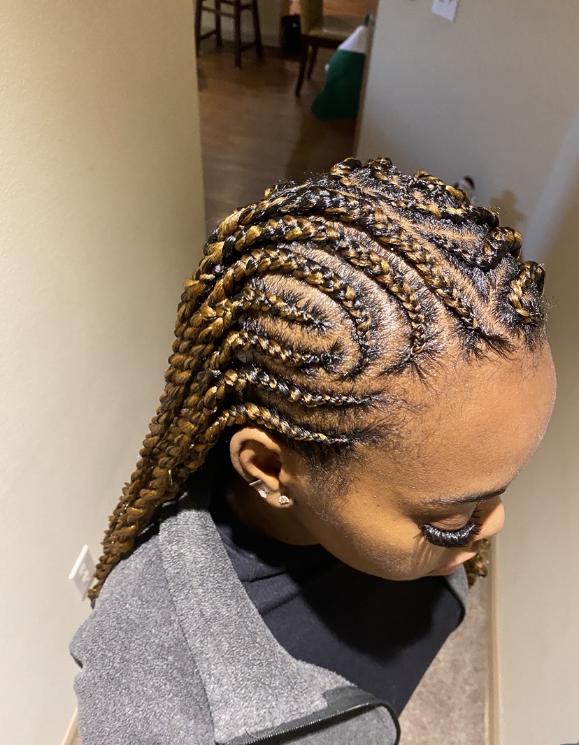 Side view of a person with intricately styled blonde and dark braids, wearing a gray and black top, indoors.