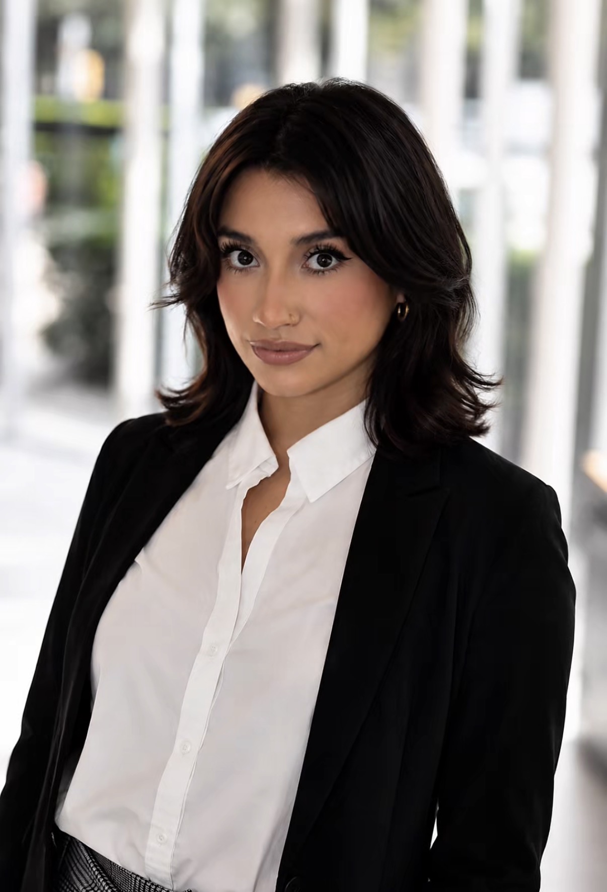 Woman with dark hair in a white shirt and black blazer, standing outdoors with blurred background.