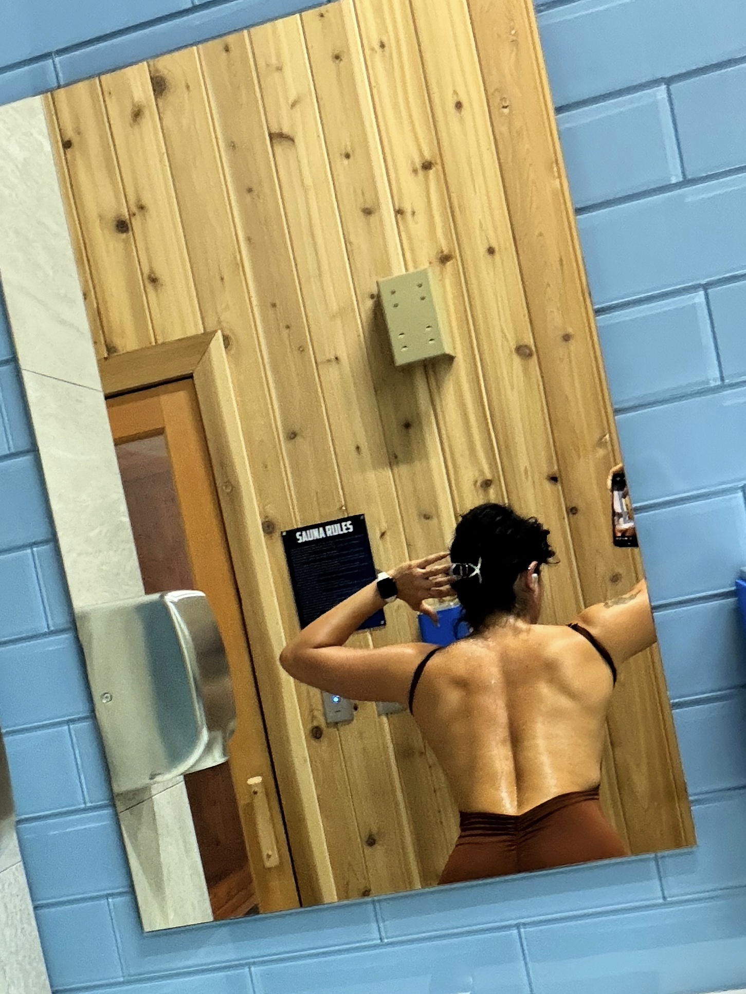 Person with short curly hair, back facing mirror, taking a selfie in a room with wooden and blue walls.