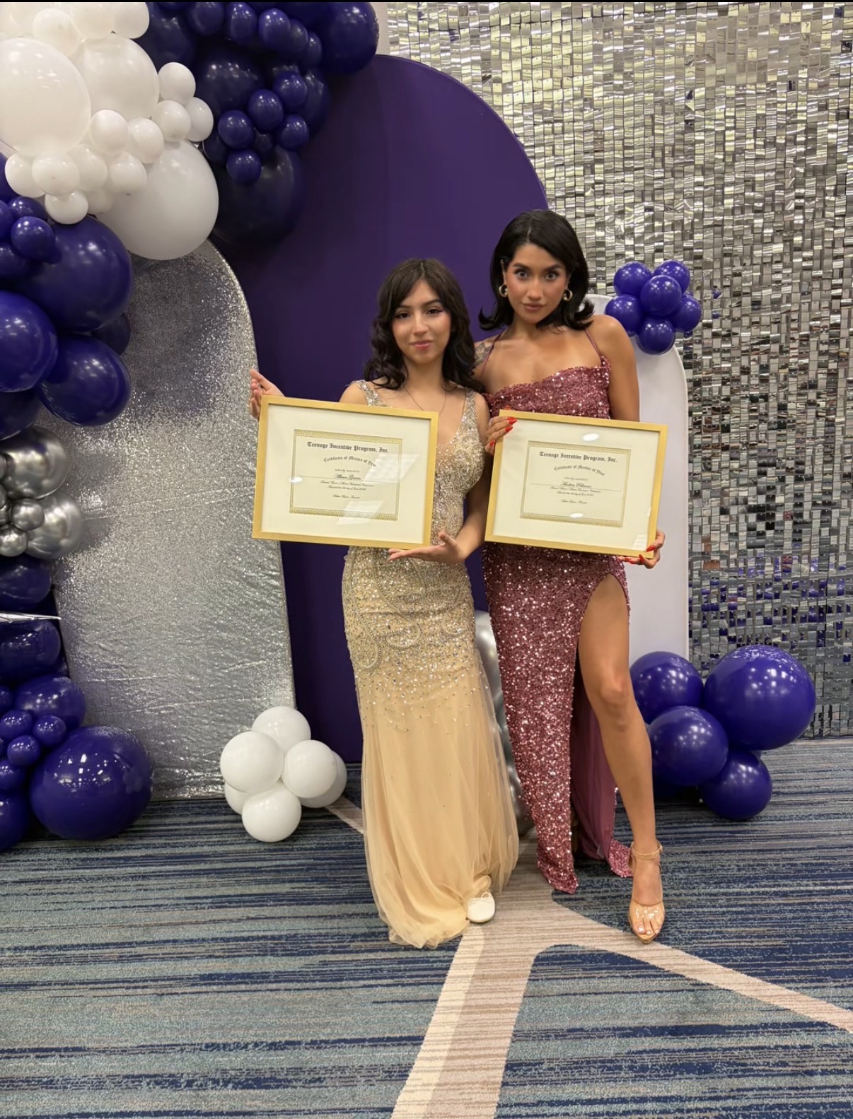 Two women in formal dresses holding certificates, standing in front of a decorated backdrop with balloons and a shiny wall.