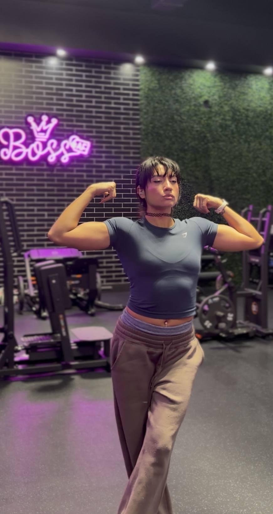 Woman in gym flexing arms, standing in front of a wall with neon sign and gym equipment, dim lighting.