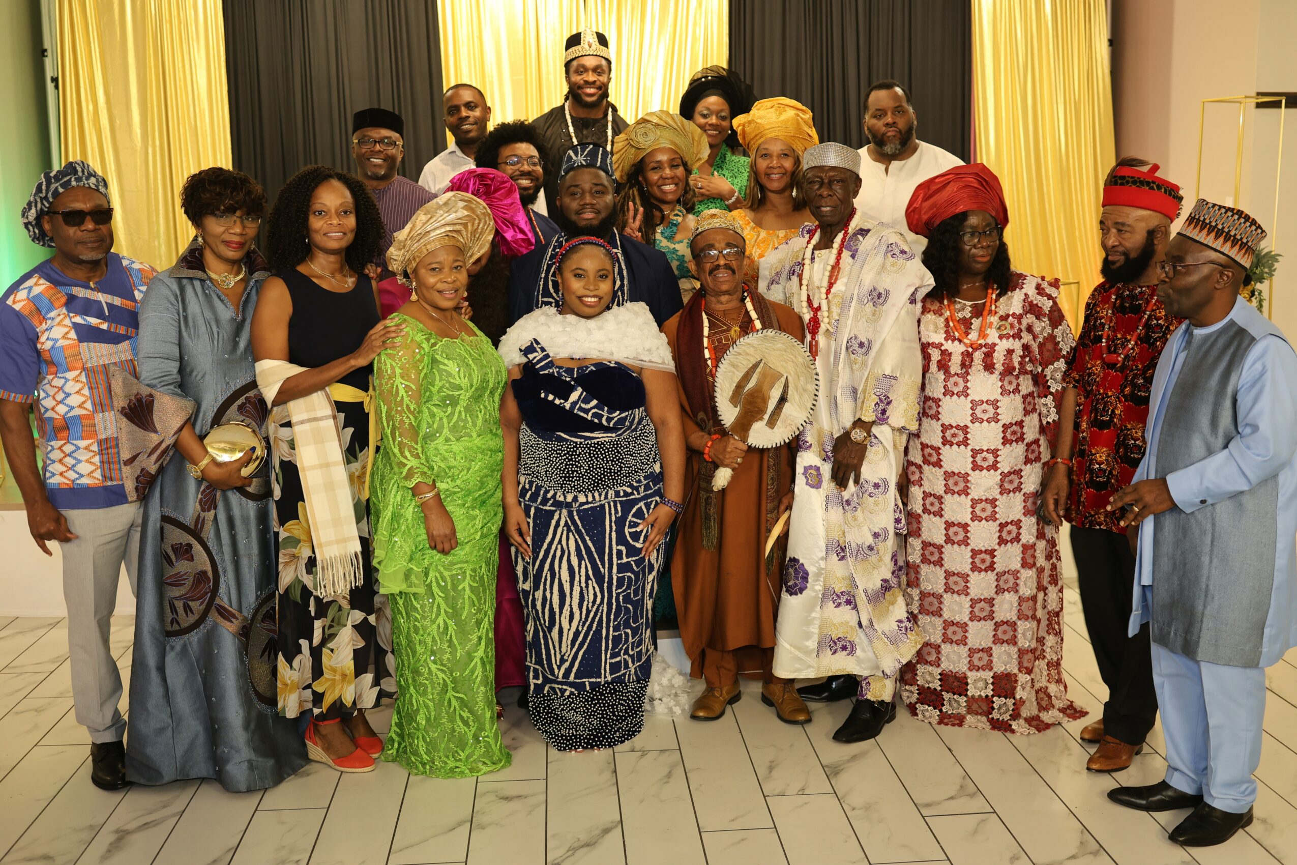 Group of people dressed in colorful traditional and modern clothing, standing indoors with yellow and black curtains behind them.