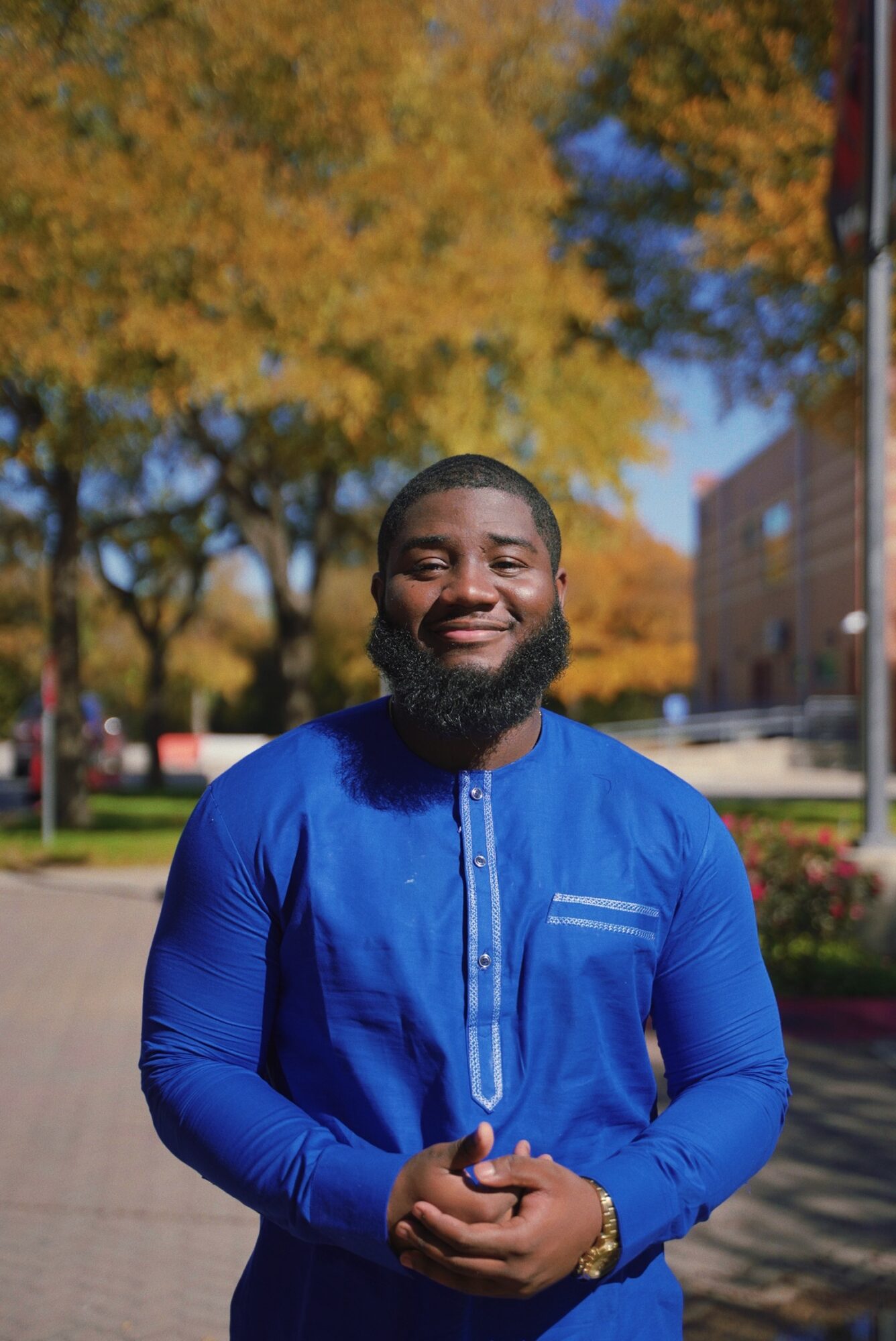 Smiling man with a beard in a blue traditional outfit outdoors with autumn trees and buildings in background.
