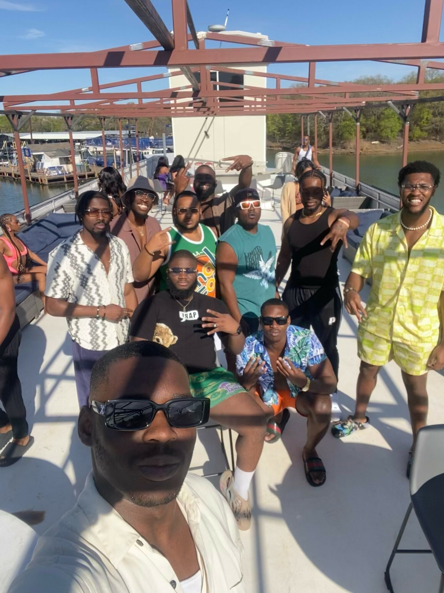 Group of people on a boat deck, some sitting and some standing, with a marina and boats in the background.