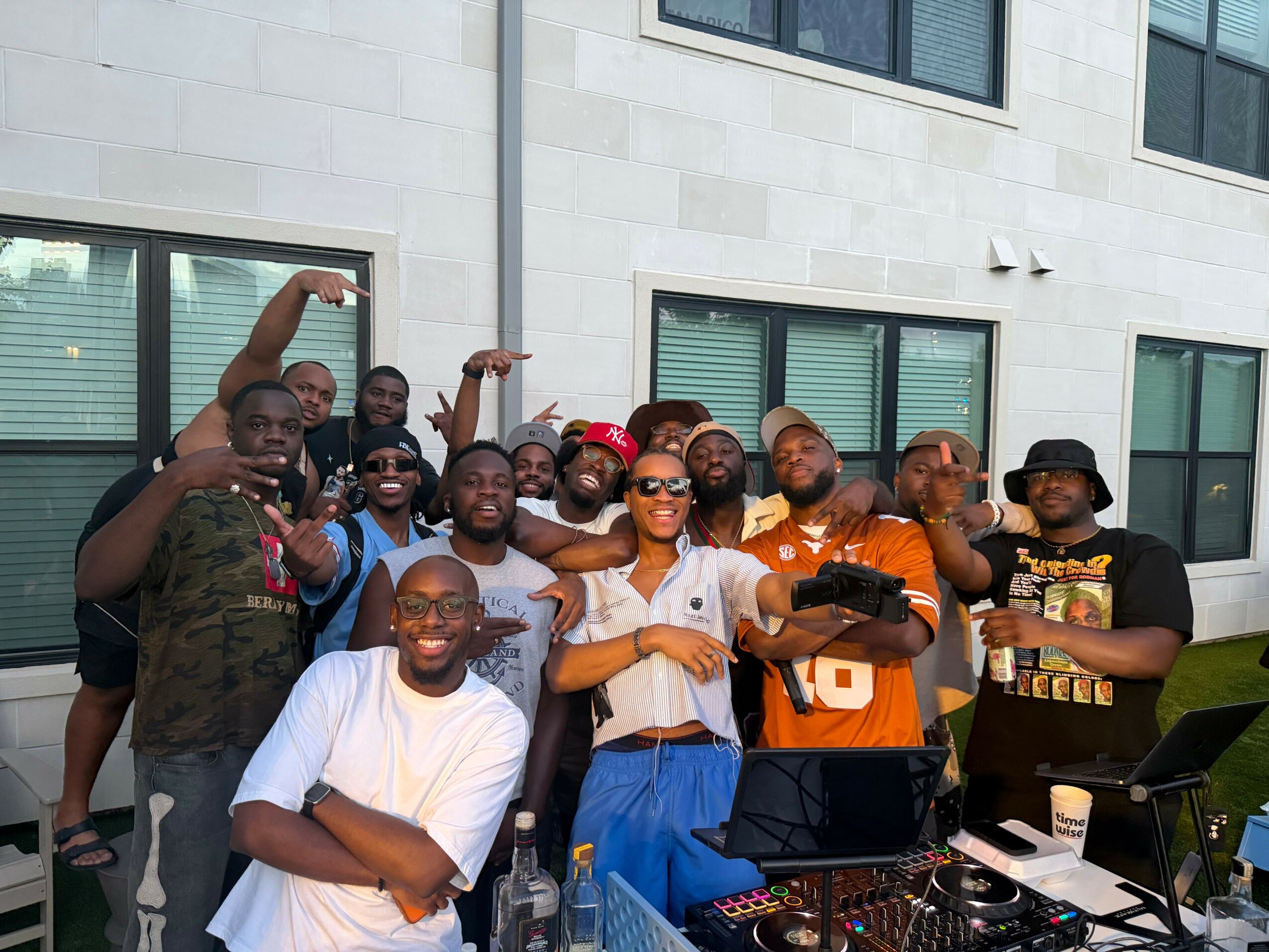 Group of people outdoors in front of a building, some holding drinks, with a DJ setup in the foreground.