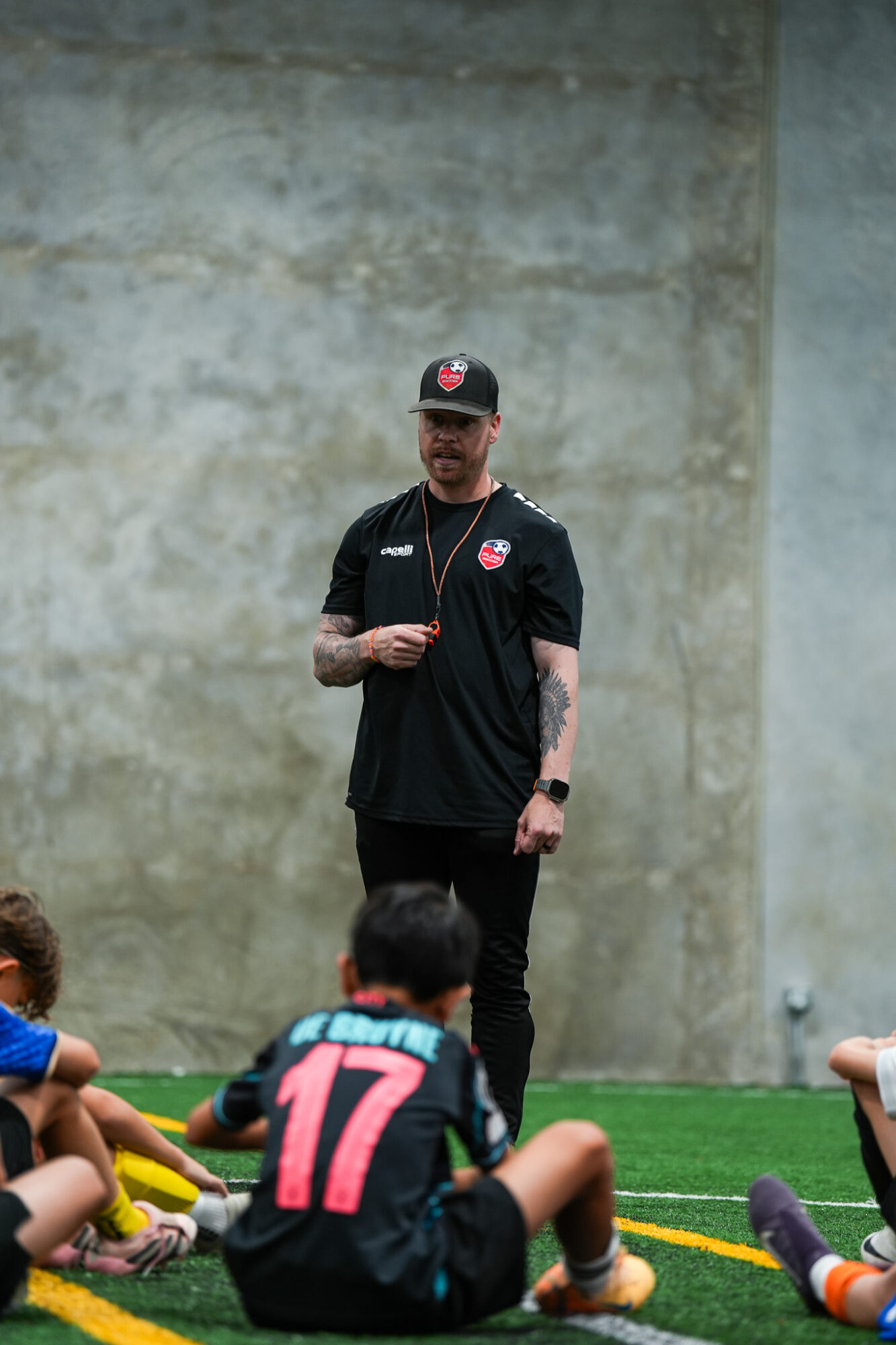 Person standing in front of a concrete wall, wearing a black sports uniform and cap, with children sitting on the ground nearby.