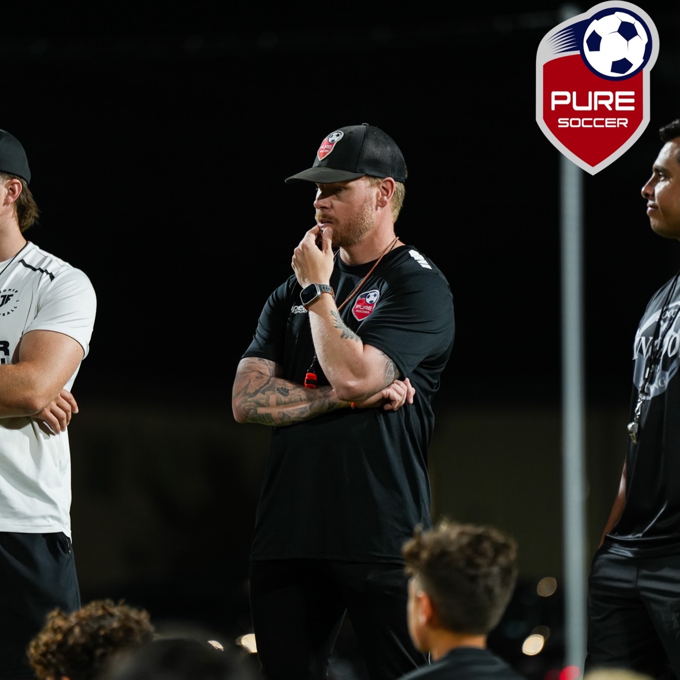 Three people standing outdoors at night, one in a black cap and shirt, another in a white shirt, and a third partially visible, near a sign that says
