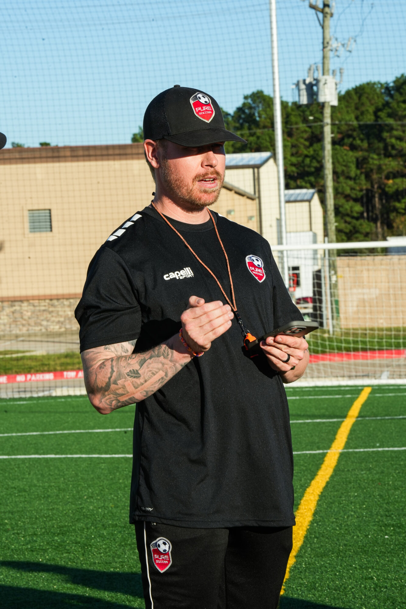 Young man with tattoos, wearing a black cap and sportswear, holding a phone on a sports field.