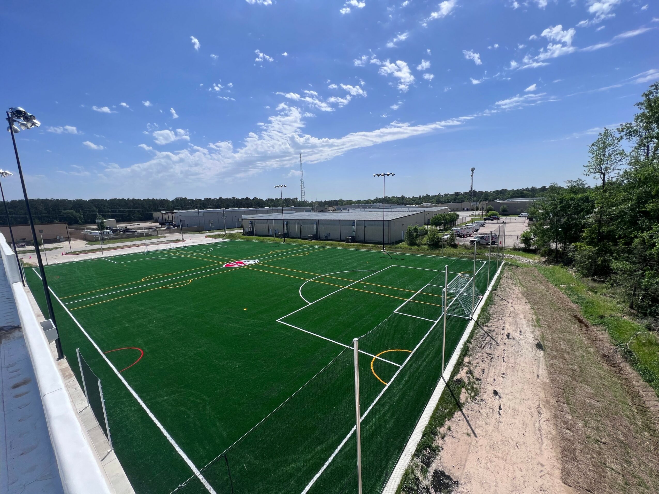 Soccer field with green artificial turf, white lines, goalposts, and surrounding fence, under a blue sky with clouds.
