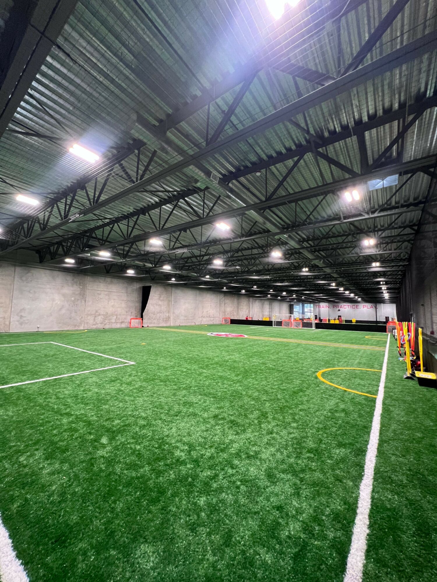 Indoor sports facility with artificial green turf, white boundary lines, and overhead lighting, viewed from the side.
