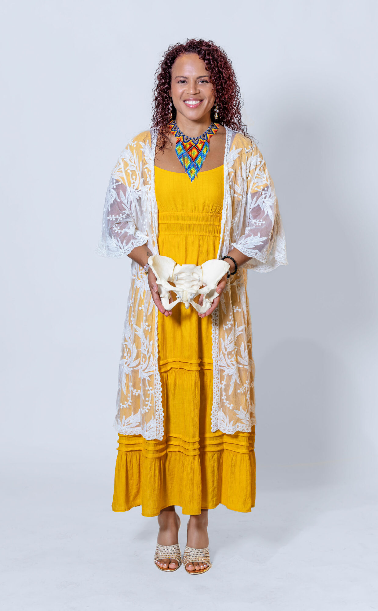 Woman in yellow dress with white lace cardigan, colorful necklace, smiling, standing against a plain background.
