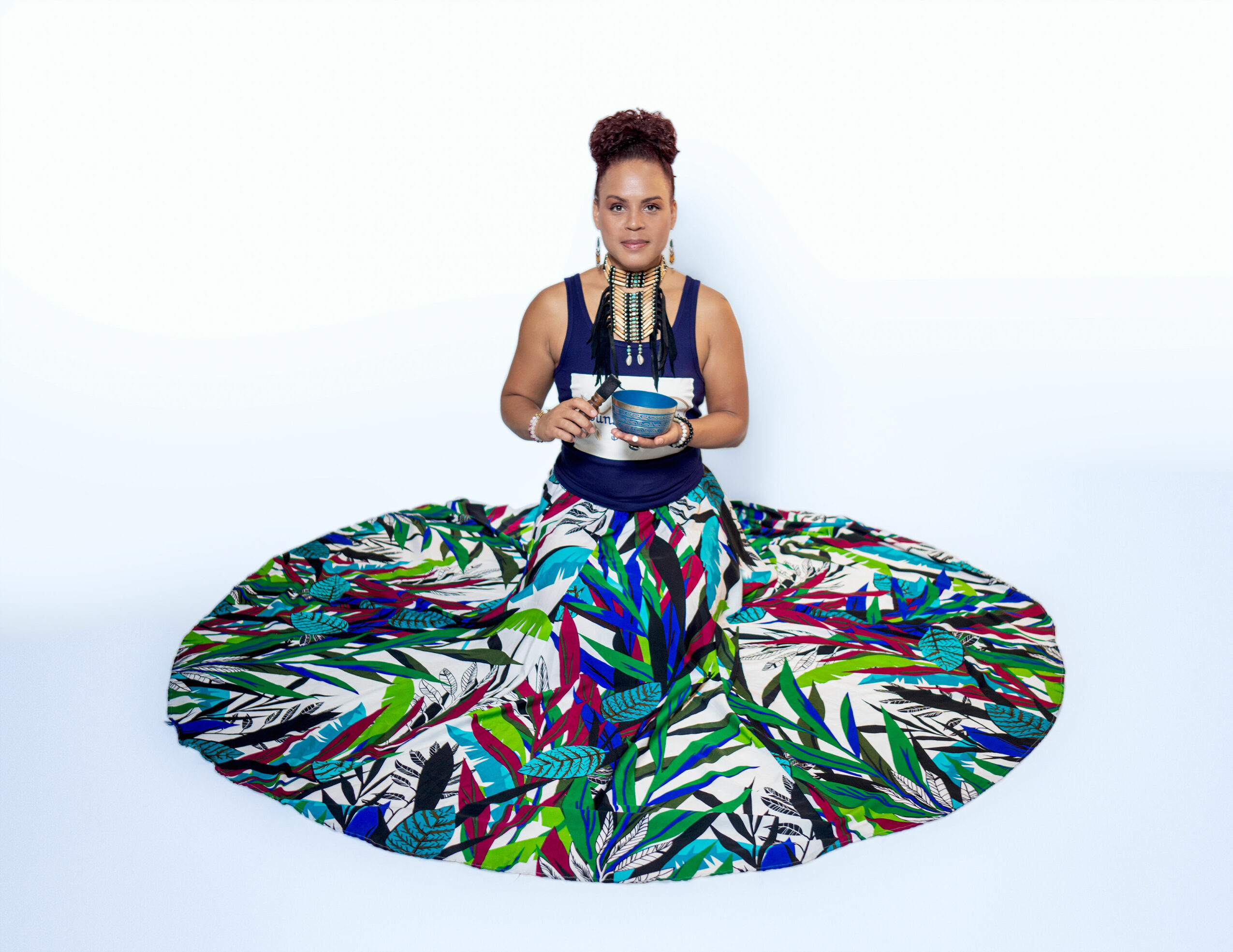 Woman sitting cross-legged on a colorful, patterned fabric, holding a bowl and spoon, against a plain white background.