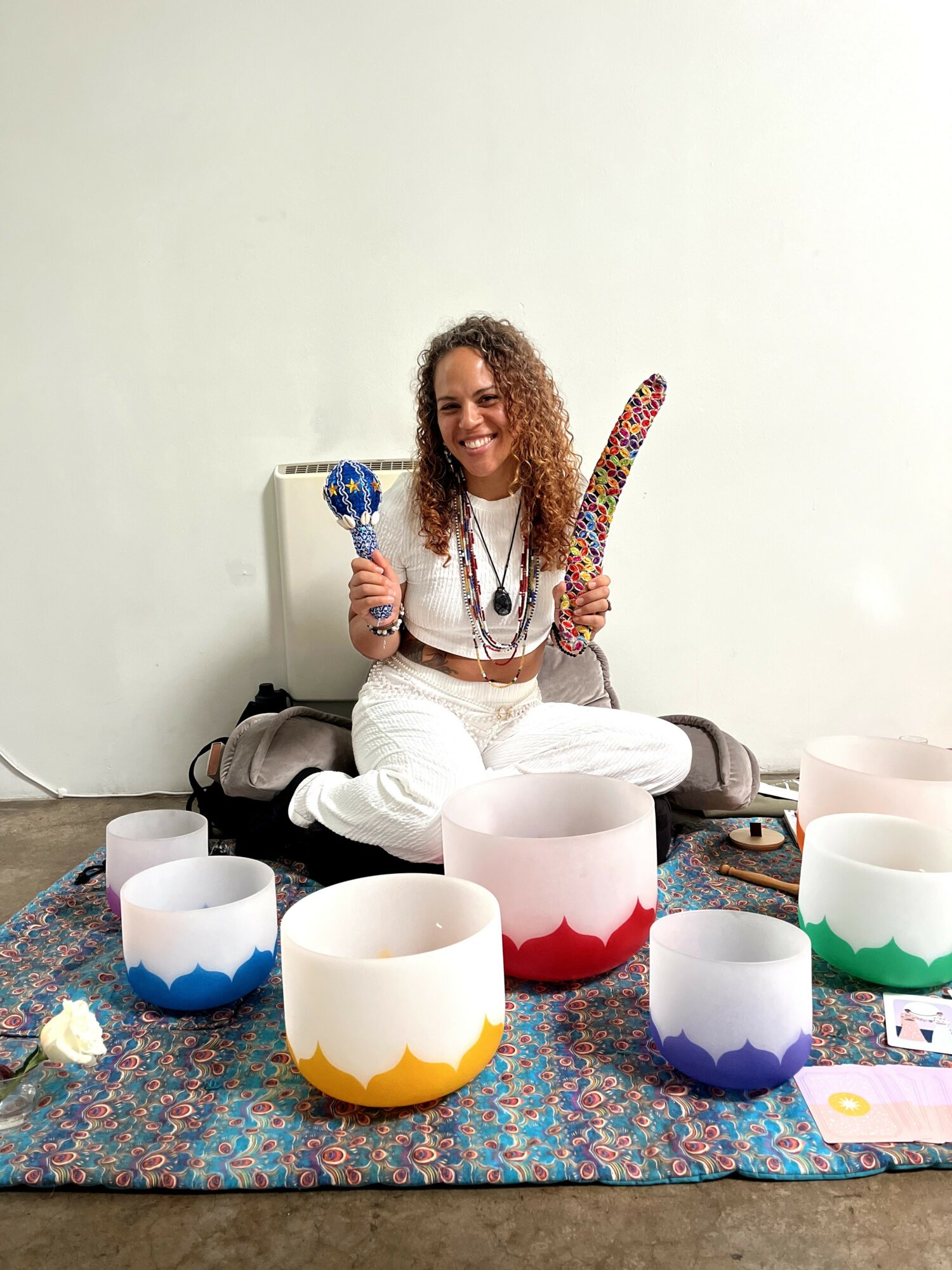 Woman sitting cross-legged on a colorful rug, holding a decorated object and a curved item, surrounded by colorful bowls.