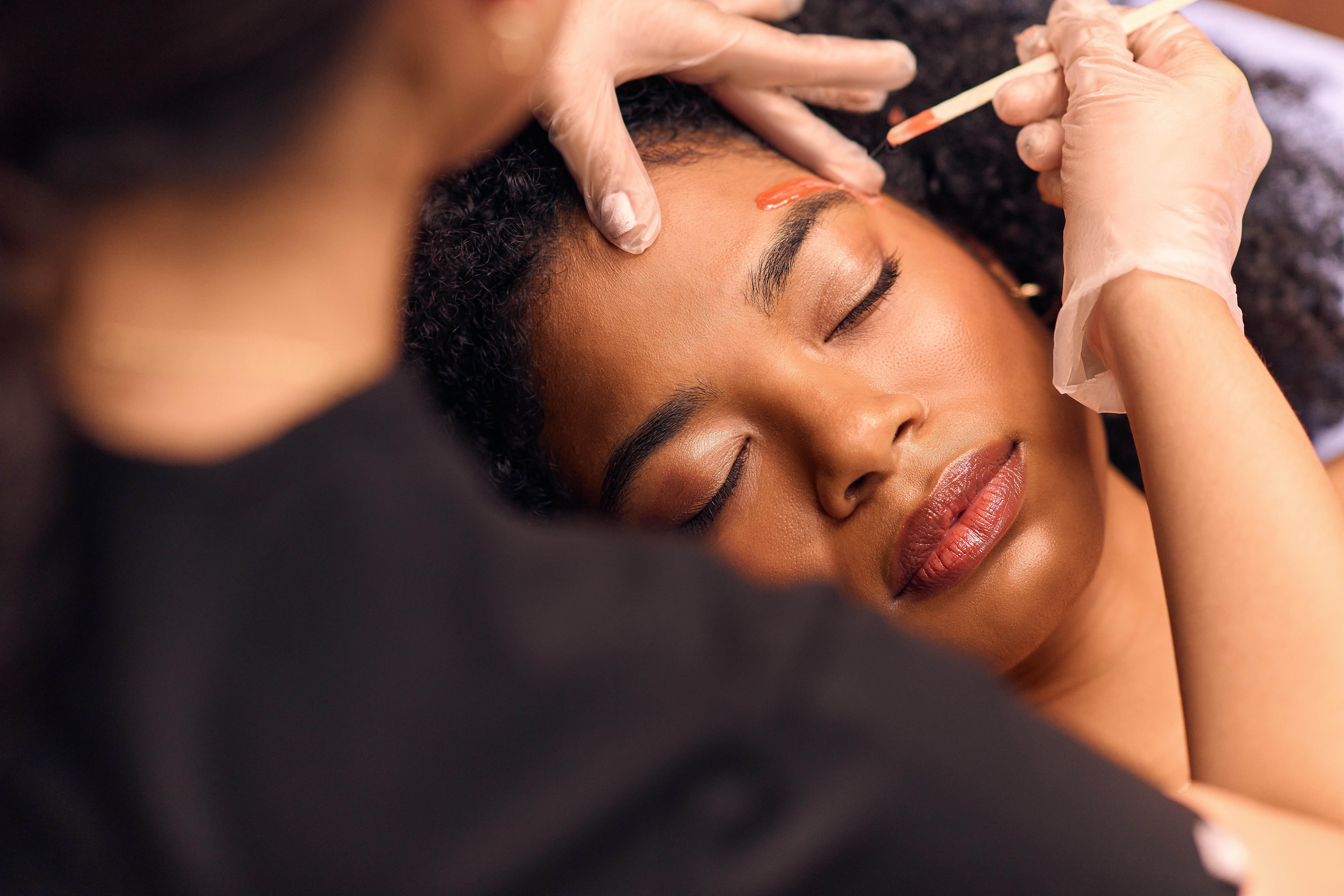 Woman receives cosmetic treatment on her face with closed eyes, person in black attire applies product with a brush, gloves worn.
