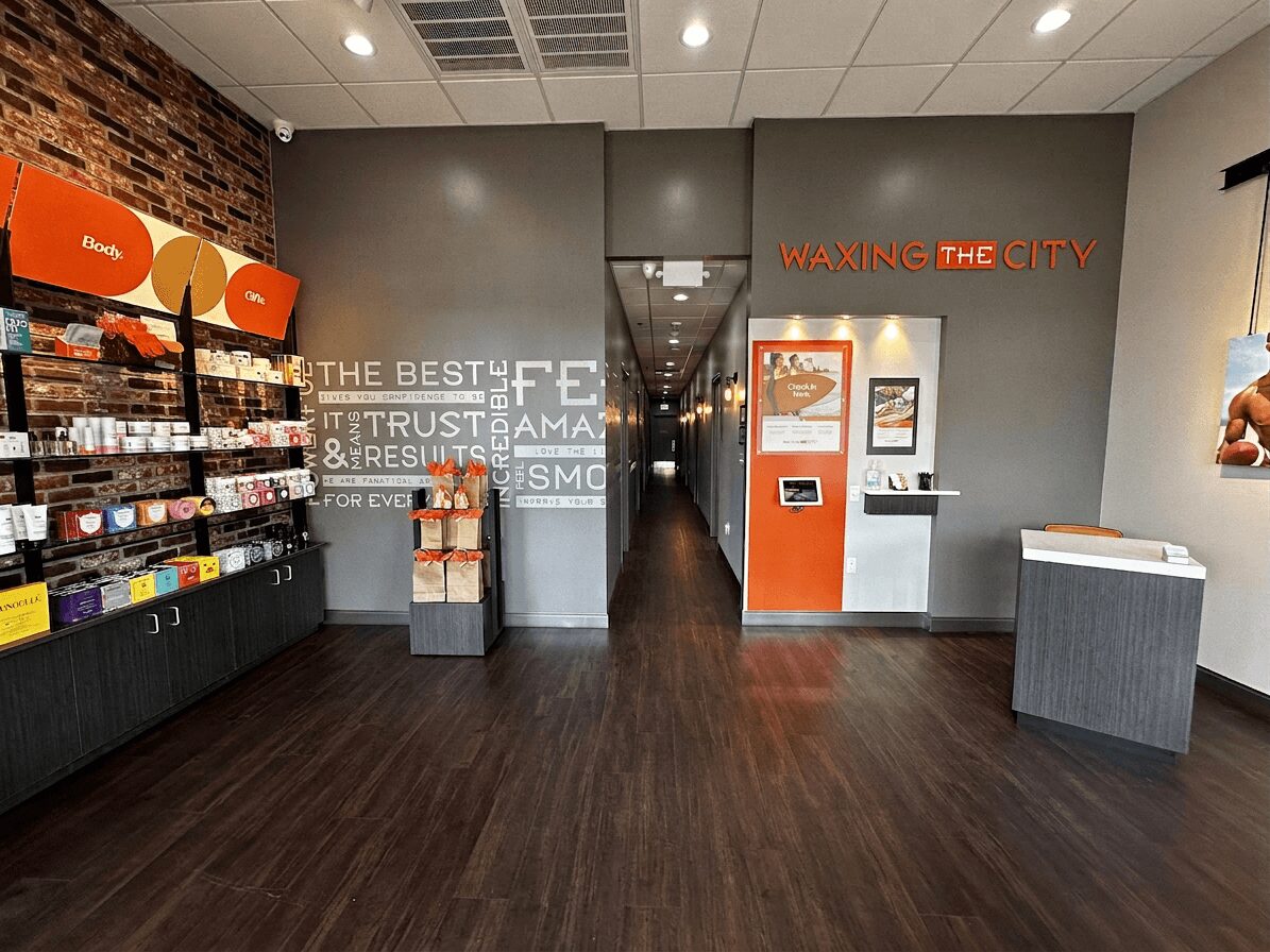 Interior of a store with shelves on the left, a gray wall with text, and an orange kiosk in the center, wooden floor.