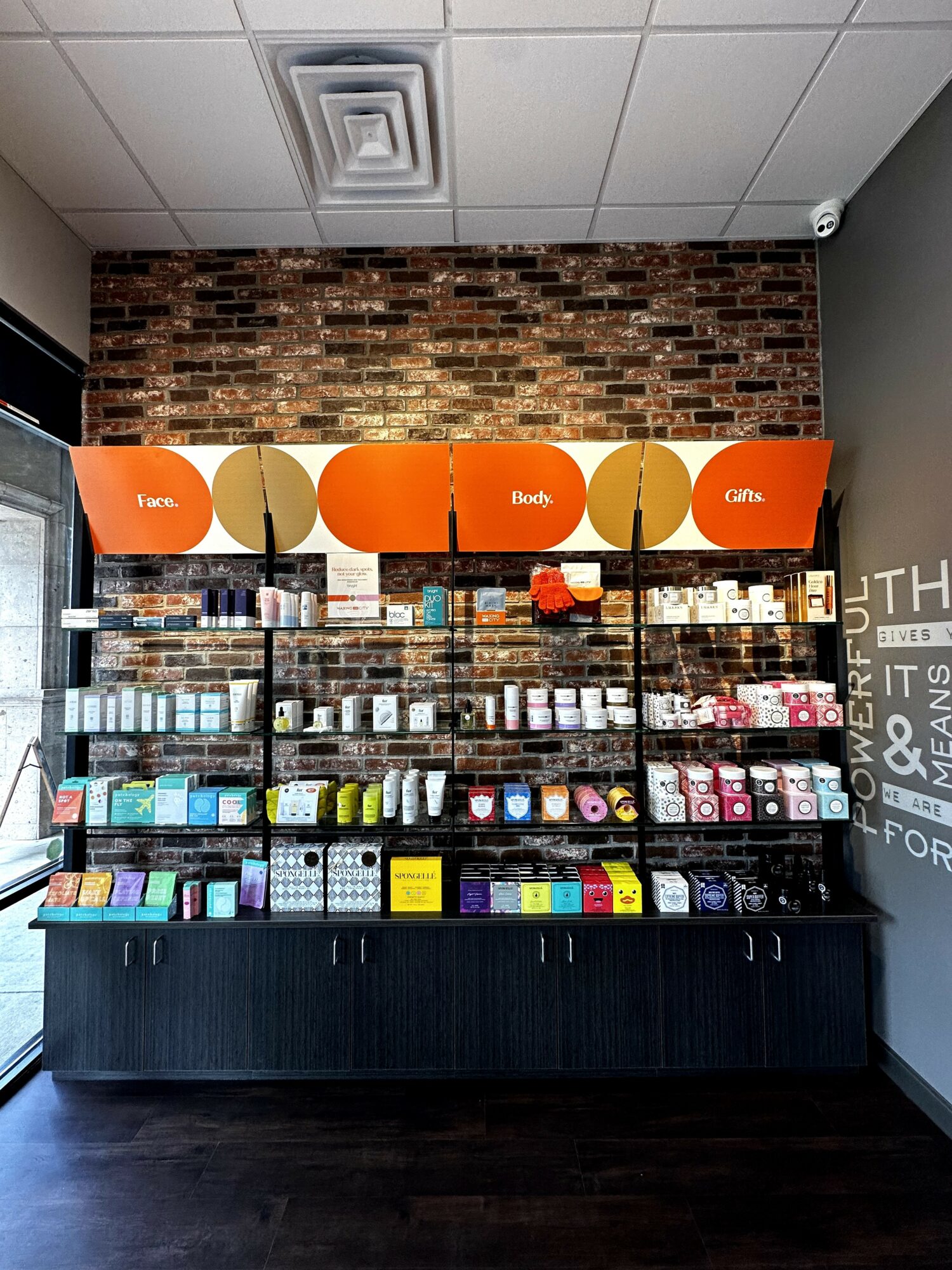 Shelves with various products in a store, orange signs labeled 'Tins,' 'Boxes,' and 'Gifts,' brick wall background.